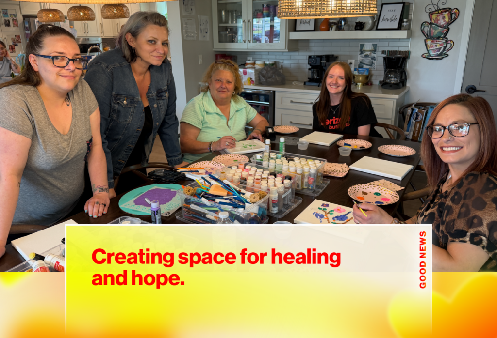 Group of five women gathered around a table with art supplies in a bright kitchen, smiling during a crafting session; banner text reads 'Creating space for healing and hope.'
