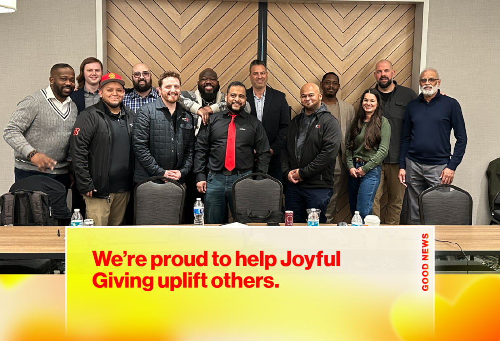 Group of diverse people standing together in a meeting room, smiling, behind a banner that reads 'We're proud to help Joyful Giving uplift others'.