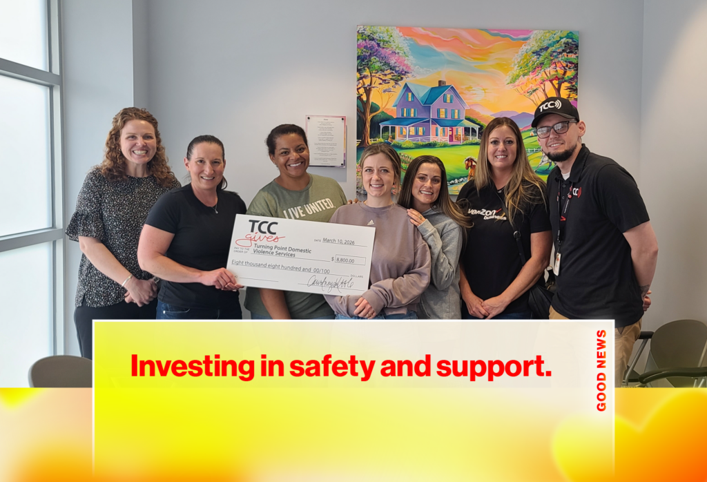Eight people pose in a bright room, holding a large donation check from TCC Gives to Turning Point Domestic Violence Services, with a colorful painting on the wall behind them and a banner reading 'Investing in safety and support' at the bottom.