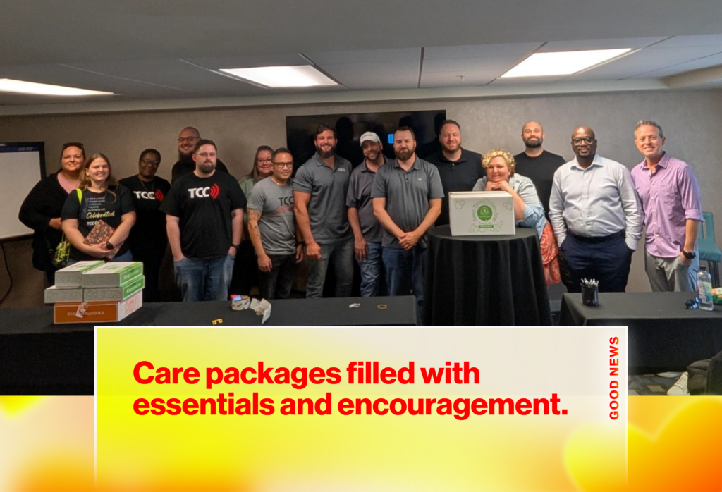 Group of coworkers posing for a team photo in a conference room with boxes on the table left side and a large screen behind them.