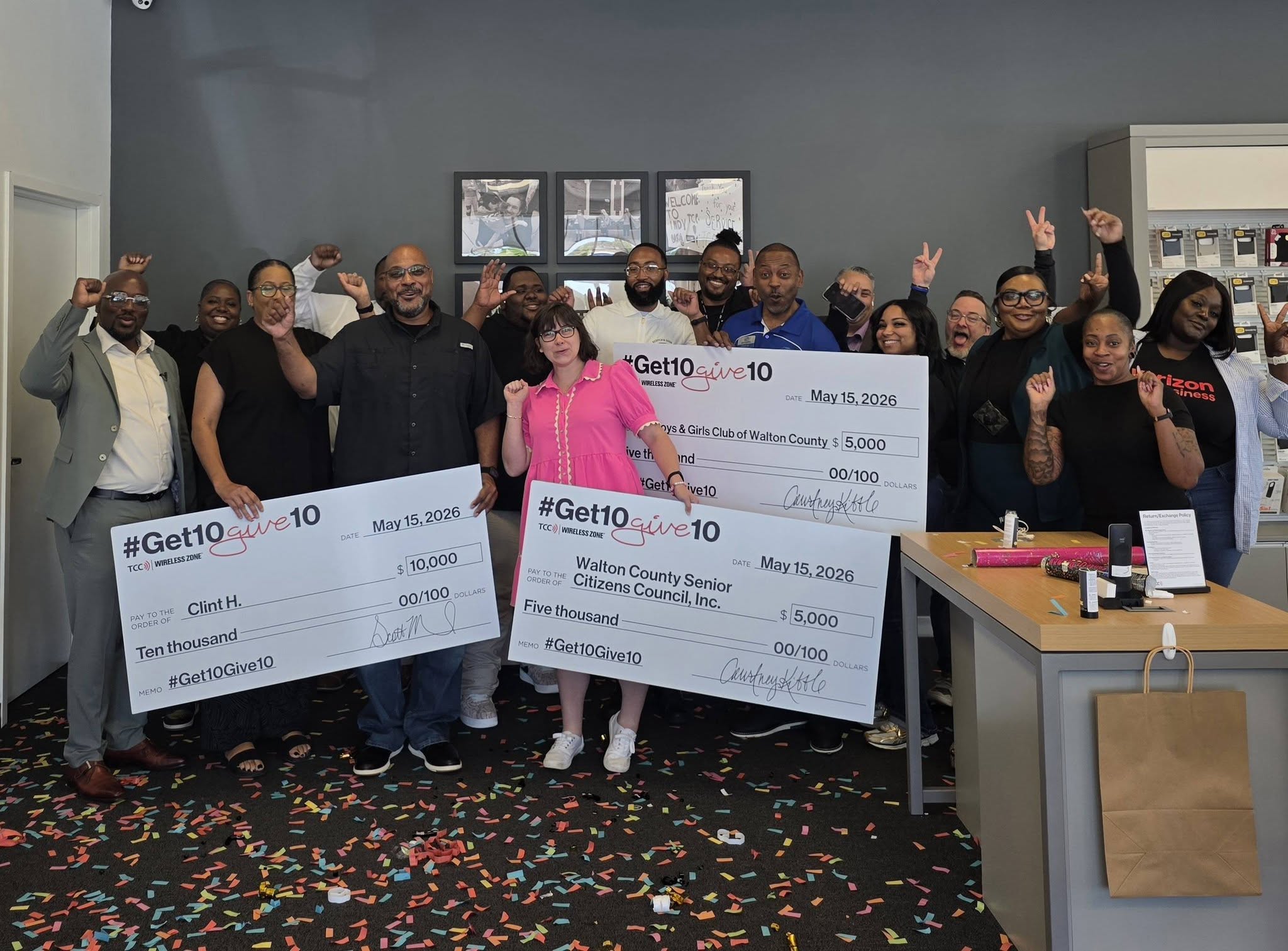 Group of people posing with oversized donation checks, celebrating a charitable gift with confetti on the floor and a desk in the background.