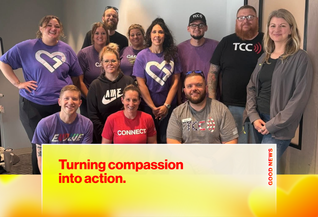 Group of coworkers posing for a team photo in a bright office, many wearing purple shirts with logos and smiling at the camera.