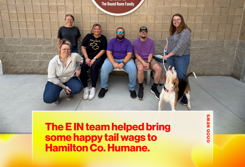 Seven people posing outside a brick building with a dog on a leash to the right and a large yellow news banner at the bottom.