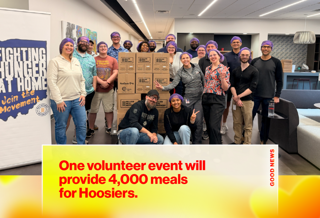Group of volunteers wearing purple hair nets posing with stacked boxes for a meal-packaging event in a community space with a hunger-relief banner nearby.