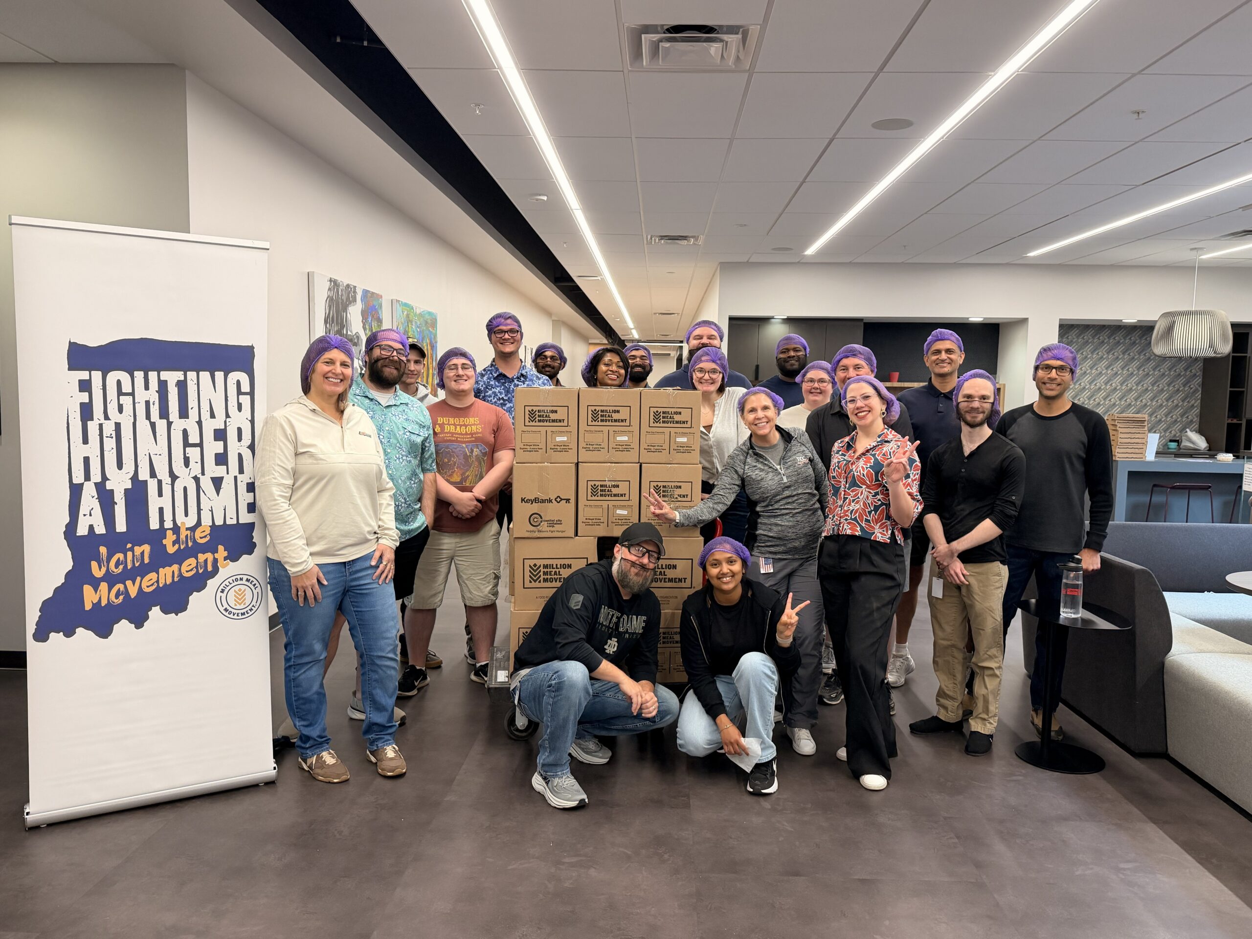 Group of volunteers wearing purple hair nets posing with stacked boxes for a hunger-relief drive in a bright office space.