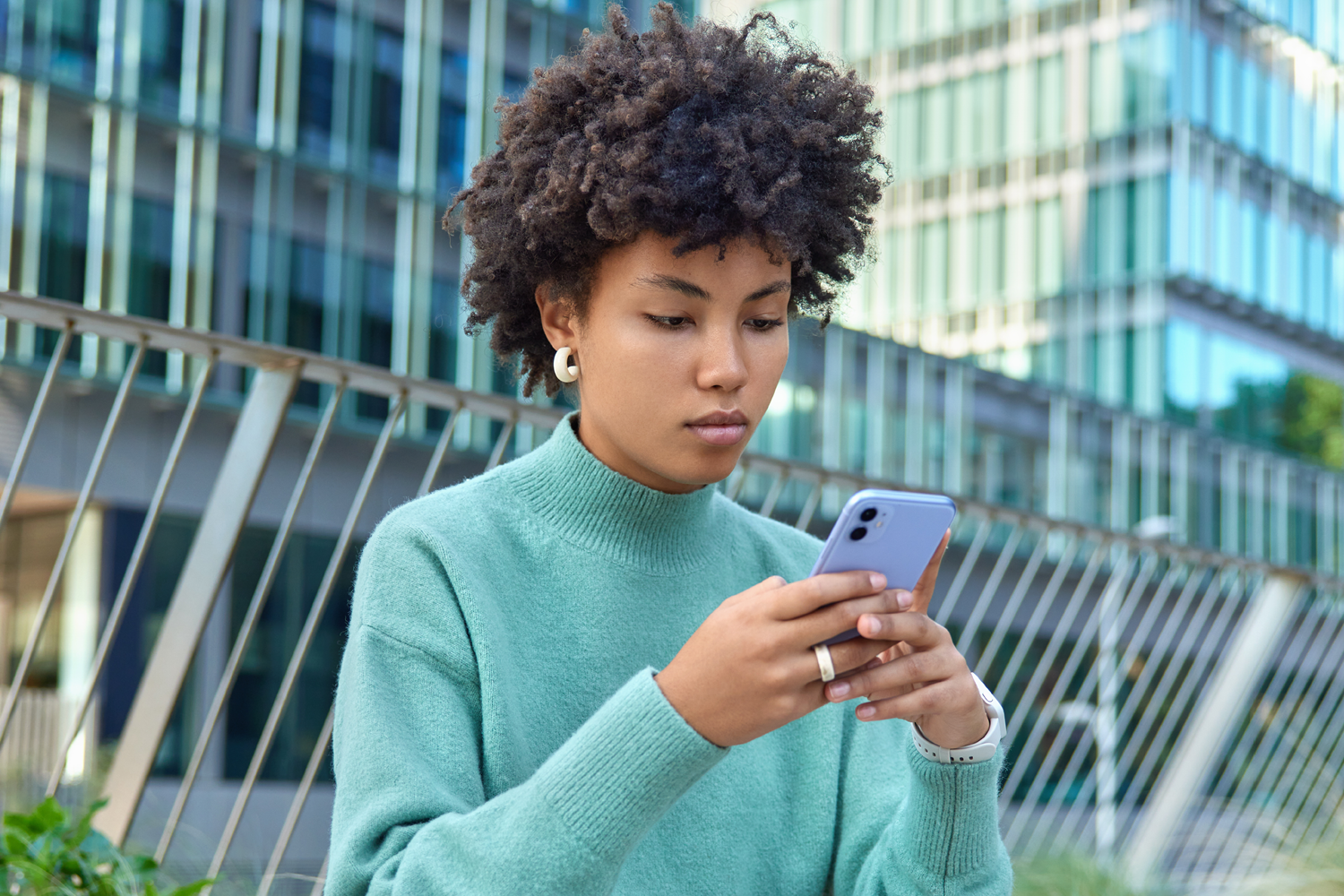 Woman with curly hair in a teal sweater checks her smartphone outdoors near a glass building with a metal railing behind her.